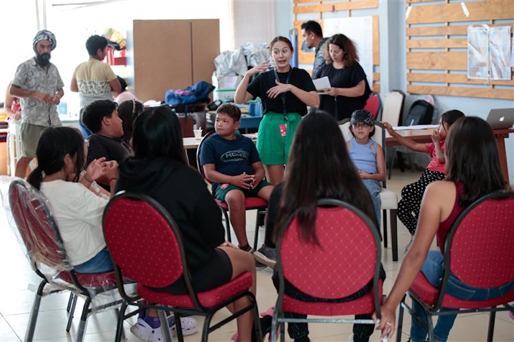 Kids and adults seated in a circle during a group activity in a room with chairs and tables.